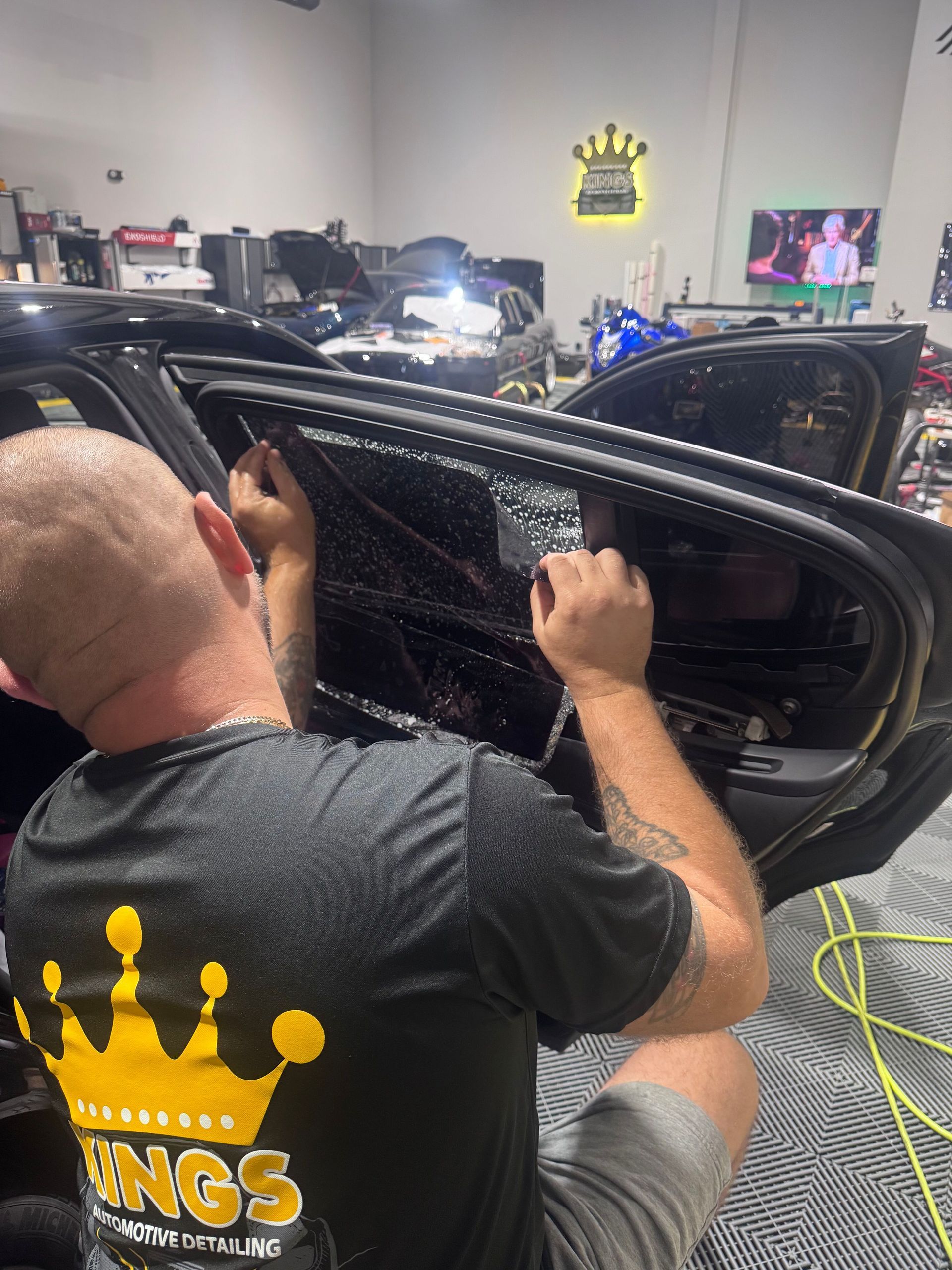 Man in a black shirt working on a car door inside a shop; a sign with a crown is on the wall.