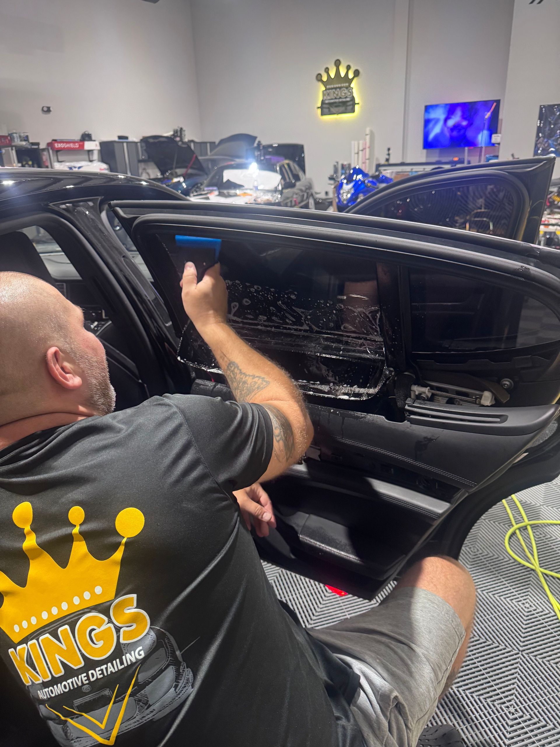 Man in black Kings shirt applying tint to a car window. Inside a shop with other cars, a tv, and a logo.