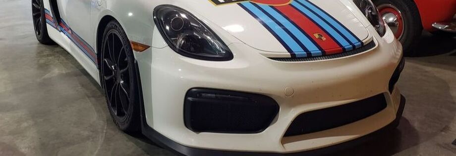 White Porsche sports car with Martini racing stripes on the hood, parked in a showroom. | Allroad Motorsports
