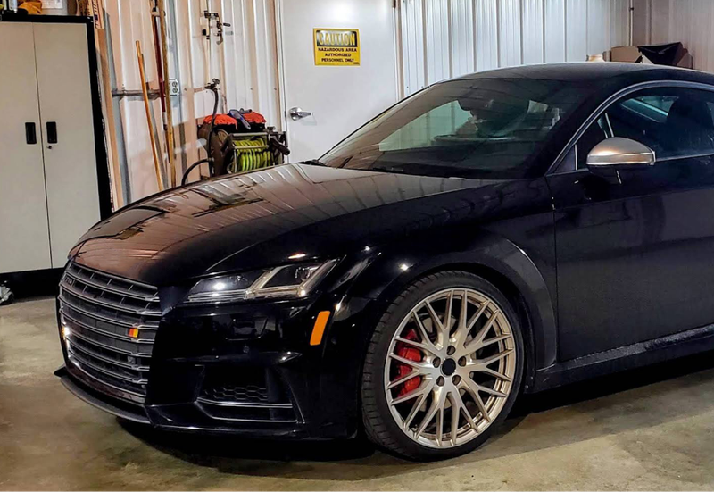 A black Audi TTS sports car parked indoors, featuring silver multi-spoke wheels with red brake calipers. | Allroad Motorsports