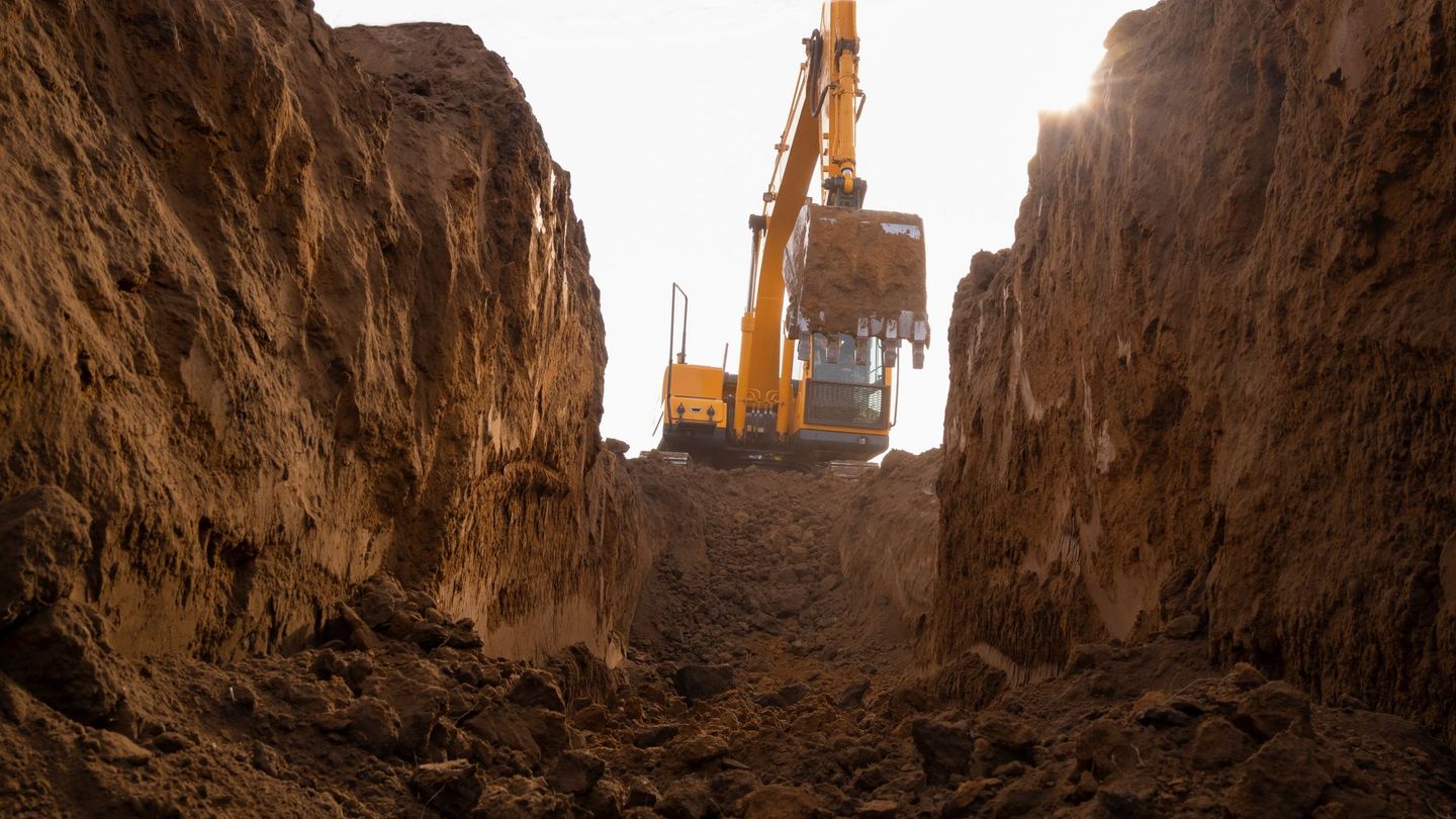Excavator digging a trench in dirt, sunlight shining from behind the machine.