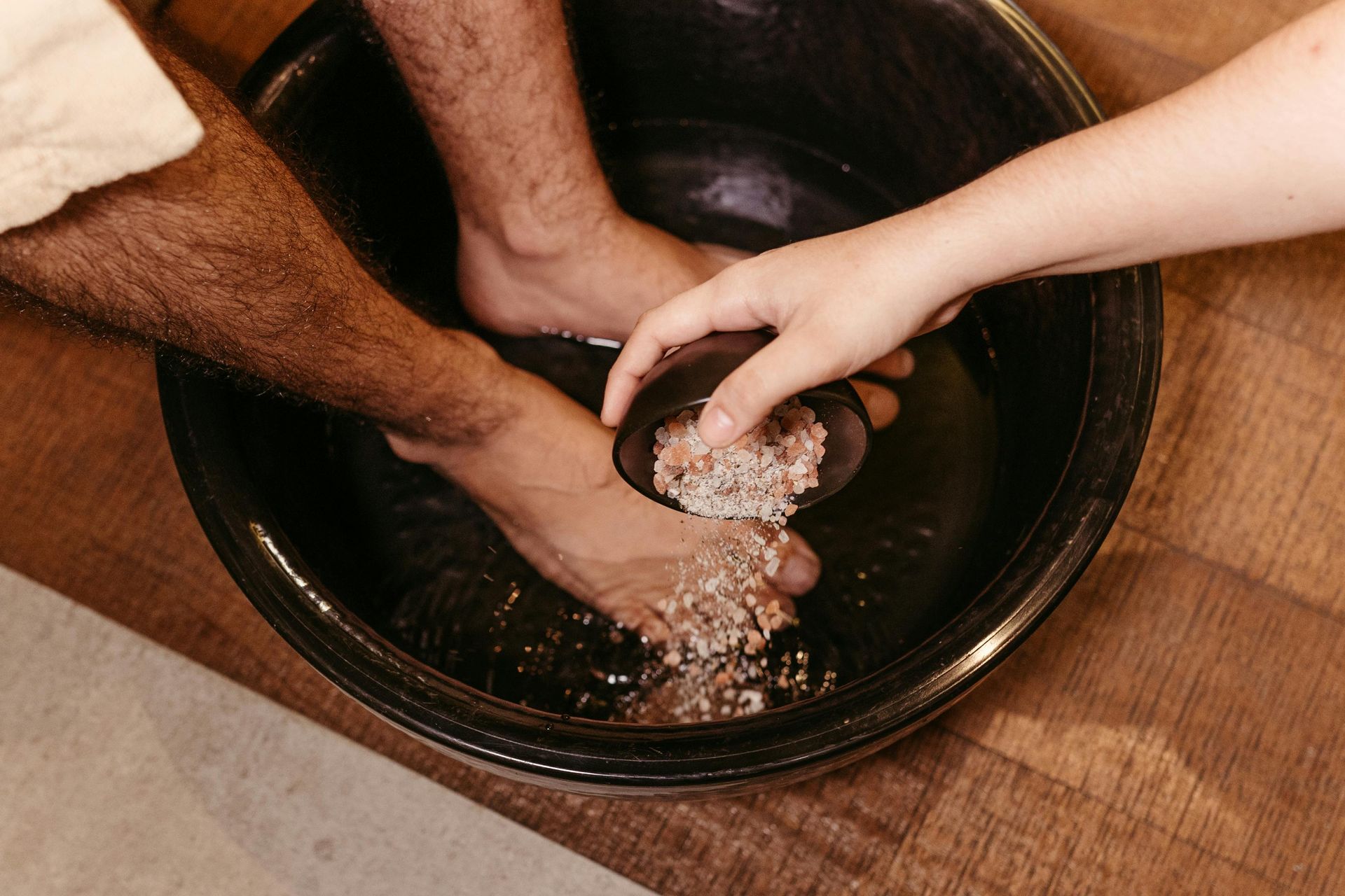 Person's feet soaking in a black basin, another person pouring salt water over the feet, indoors.