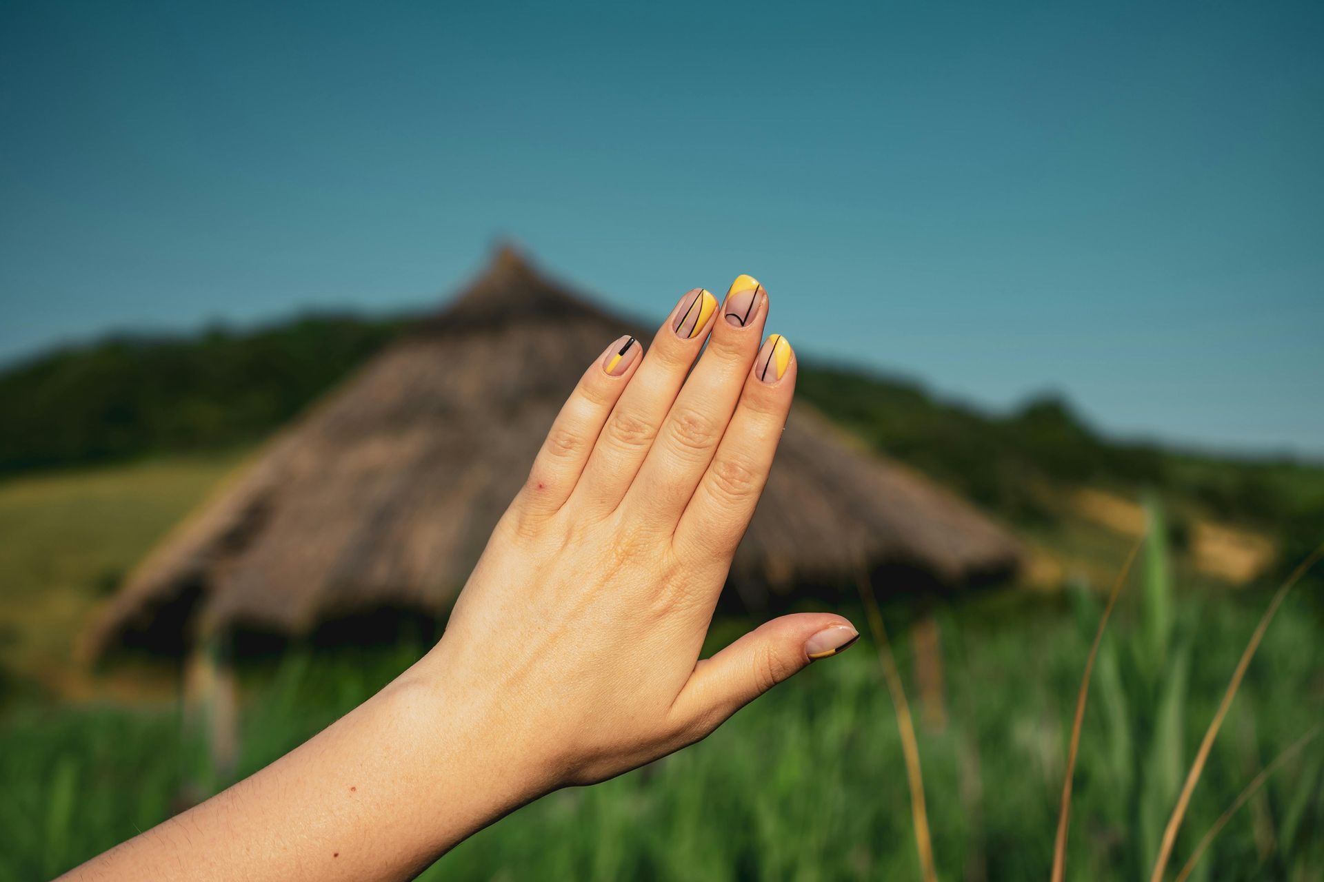 Hand with yellow and black painted nails in front of a thatched roof hut, tall grass, and blue sky.