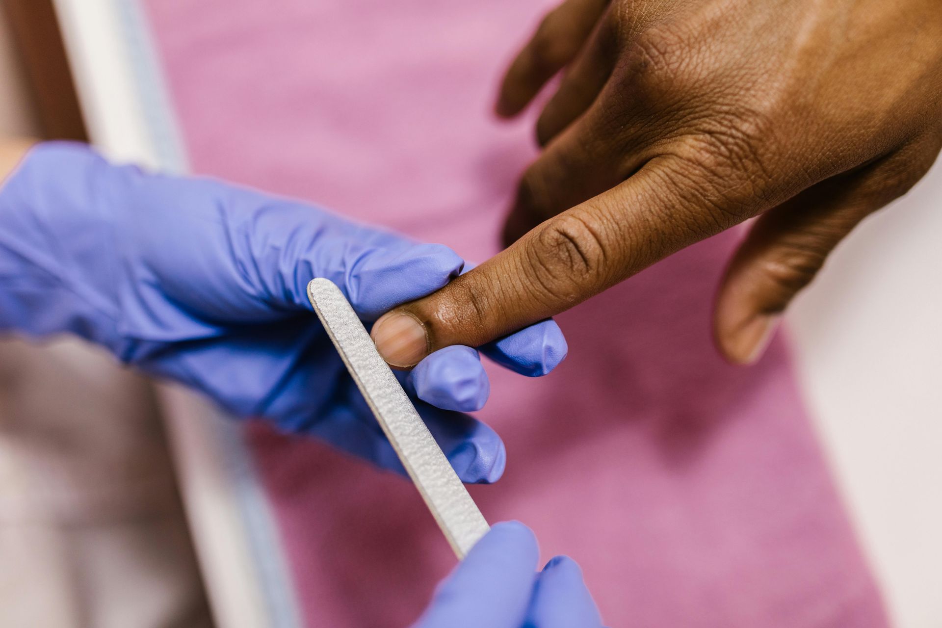 Person's fingernail being filed by gloved hands, in a salon setting, on a pink surface.