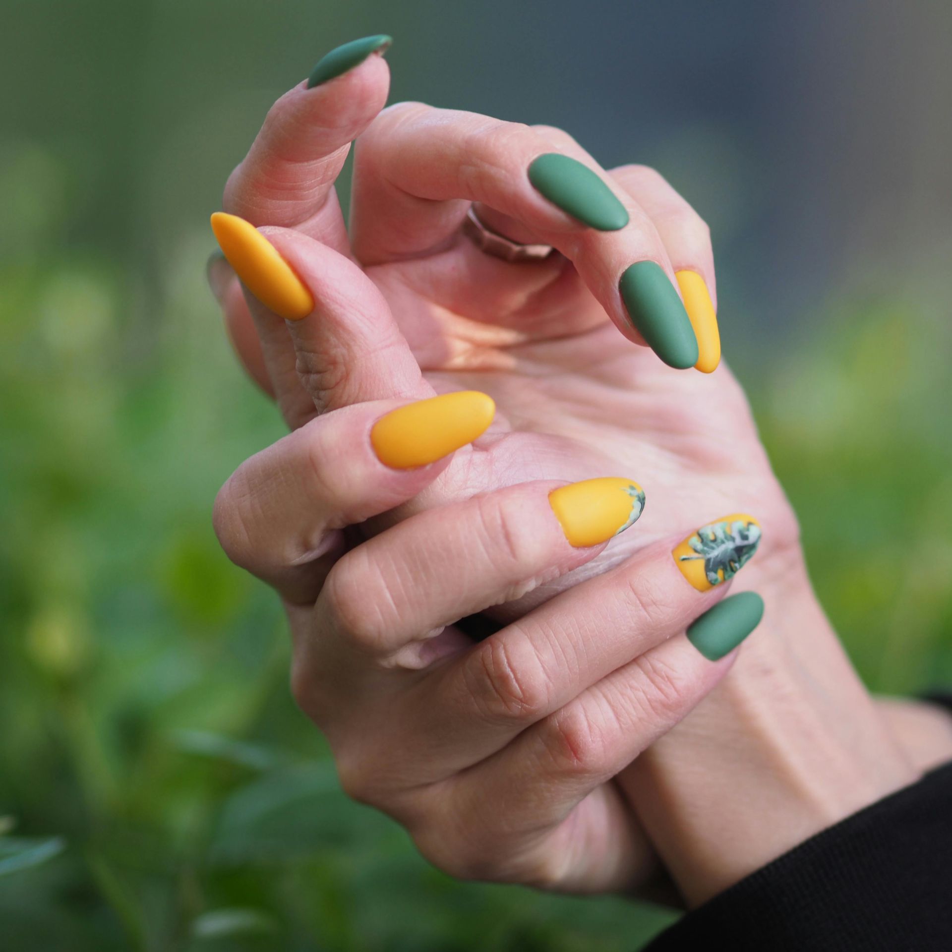 Hands with almond-shaped nails painted in matte yellow, green, and a floral design against a blurred green background.