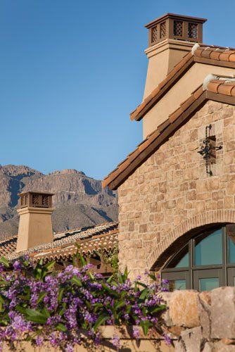 Stone house with tile roof, arched window, chimneys, and purple flowers against a mountain backdrop.