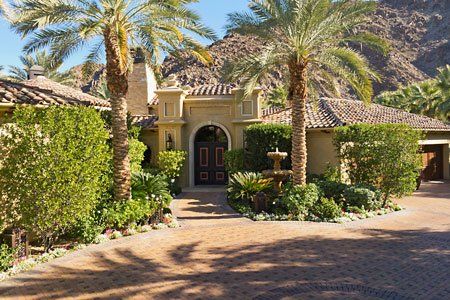 A beige stucco house with a red tiled roof, palm trees, a fountain, and a mountain backdrop.