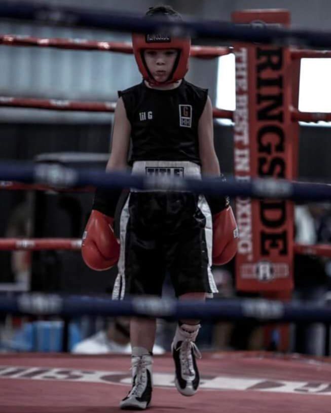 A person with braided hair poses in a boxing gym wearing a white tank top, black shorts, and black and gold boxing gloves.