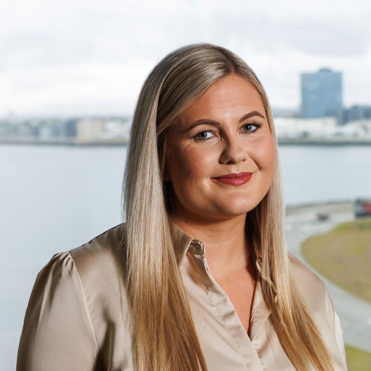 Kona með sítt ljóst hár brosir fyrir myndavélinni fyrir framan vatnið.
