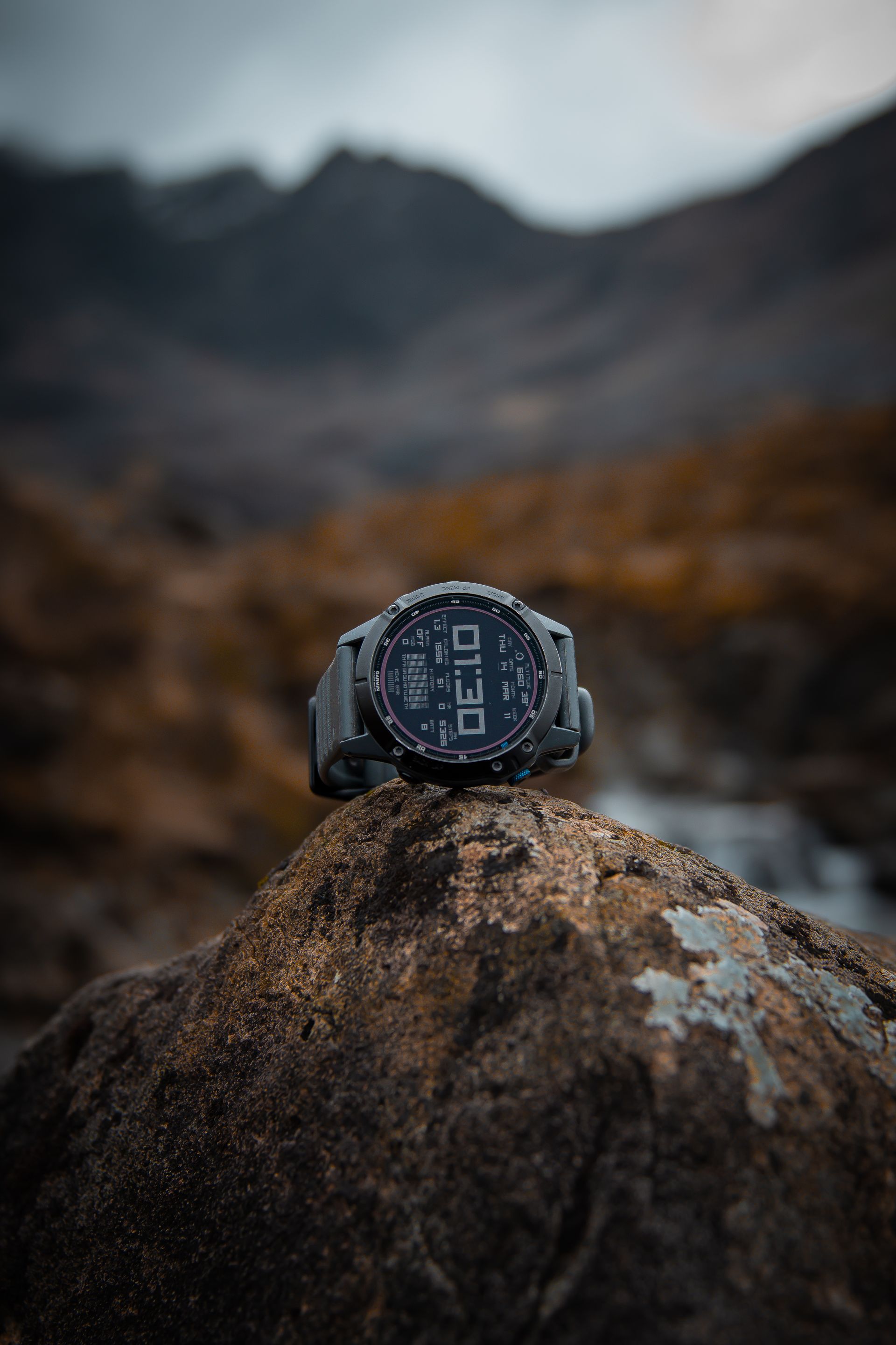 Gray smartwatch on a textured rock with a mountain range in the background.