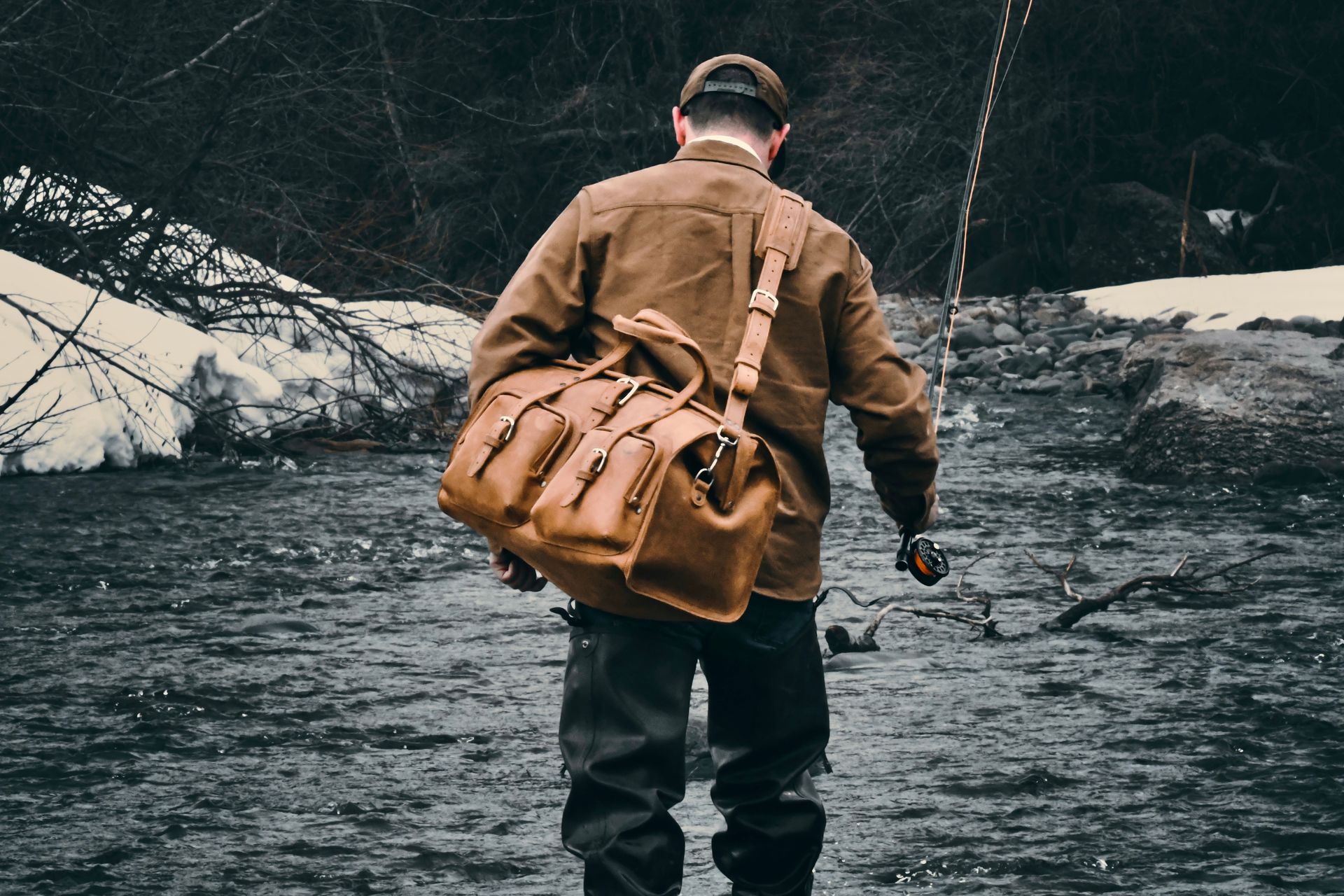 Man wading in river, fishing, with brown leather bag, wearing cap and jacket in a winter scene.