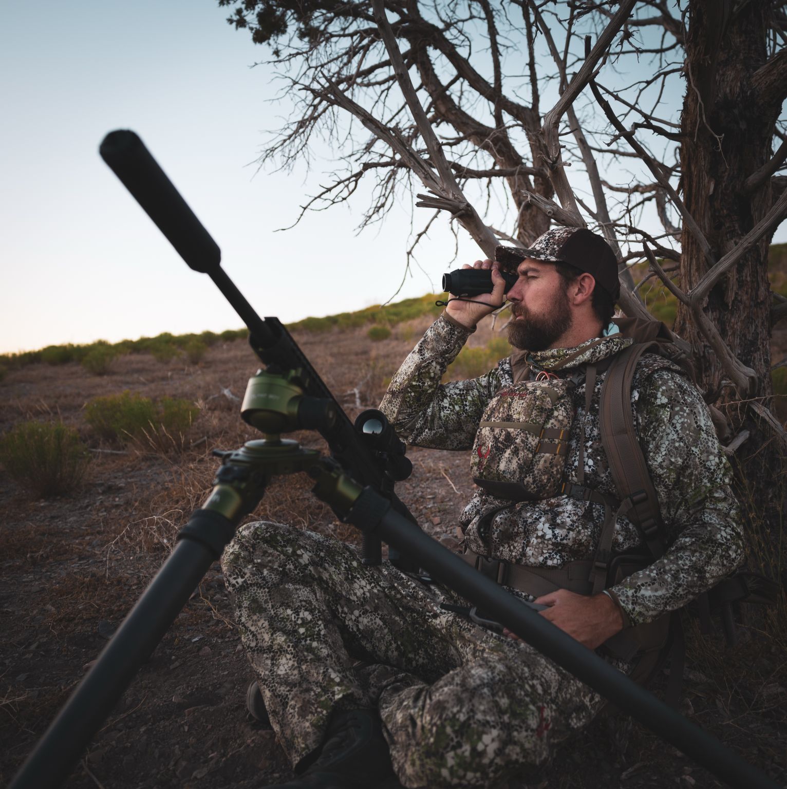 Person wearing camouflage sitting against a tree in a field.