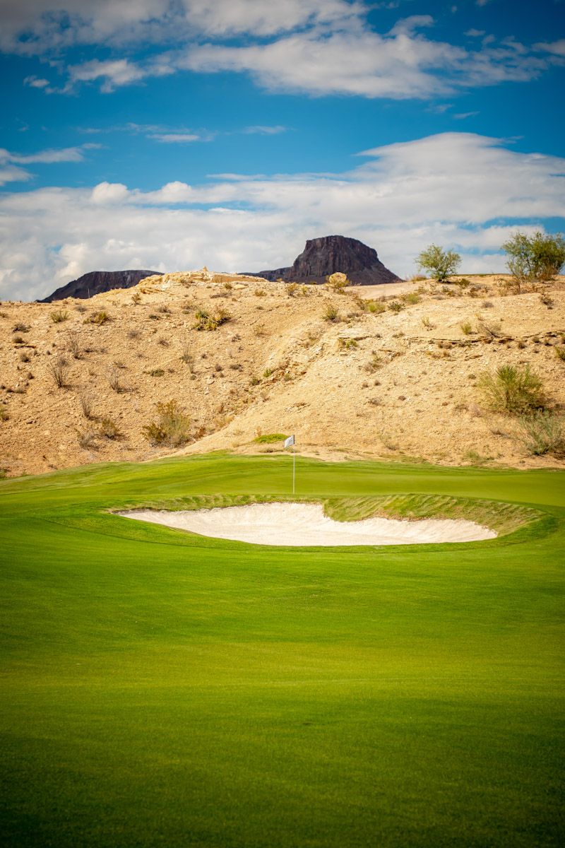 Golf green with a sand trap, flag, and a mountain backdrop under a bright blue sky.