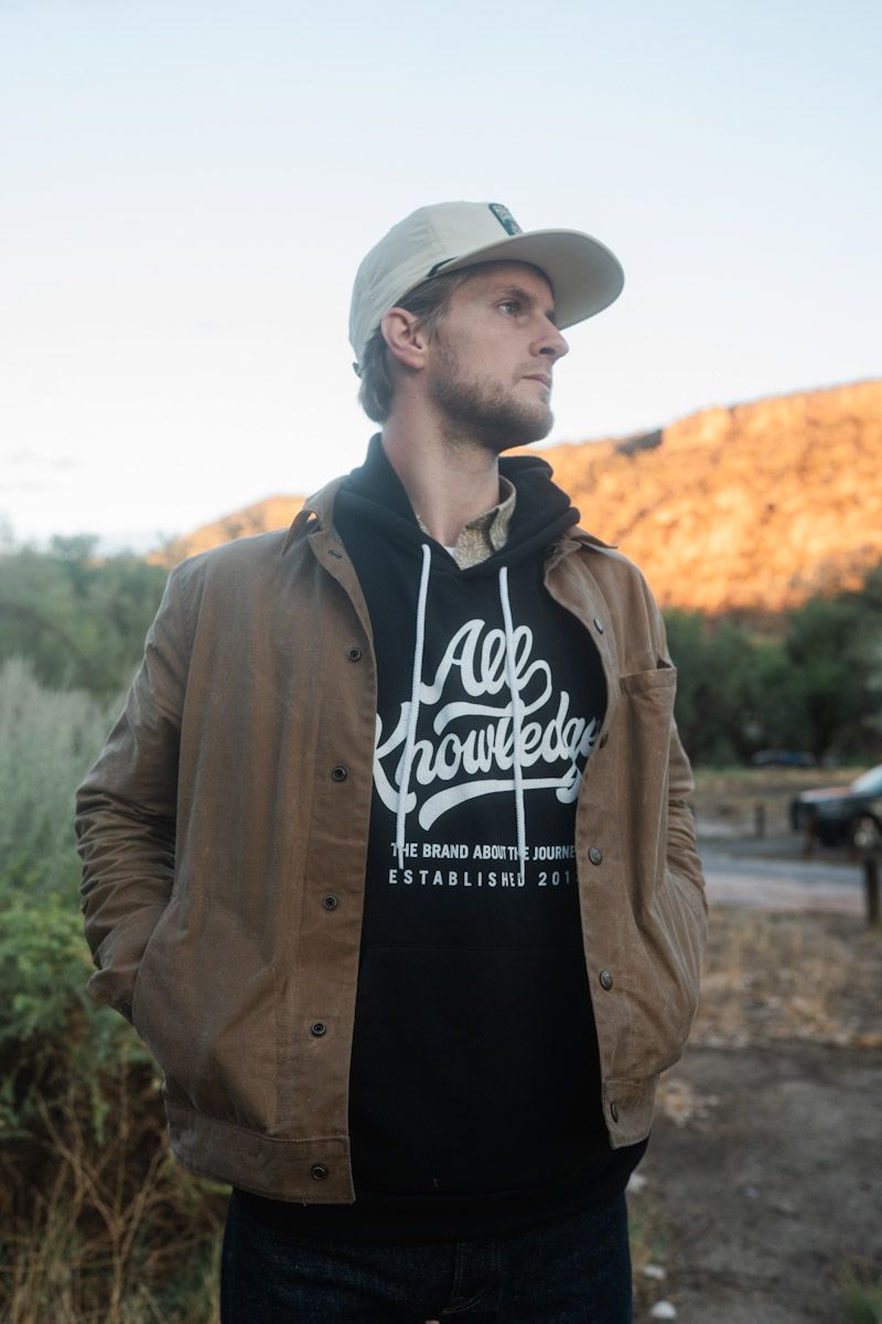 Man in jacket and cap looking off-camera, wearing a black hoodie with white lettering; outdoors in natural setting.