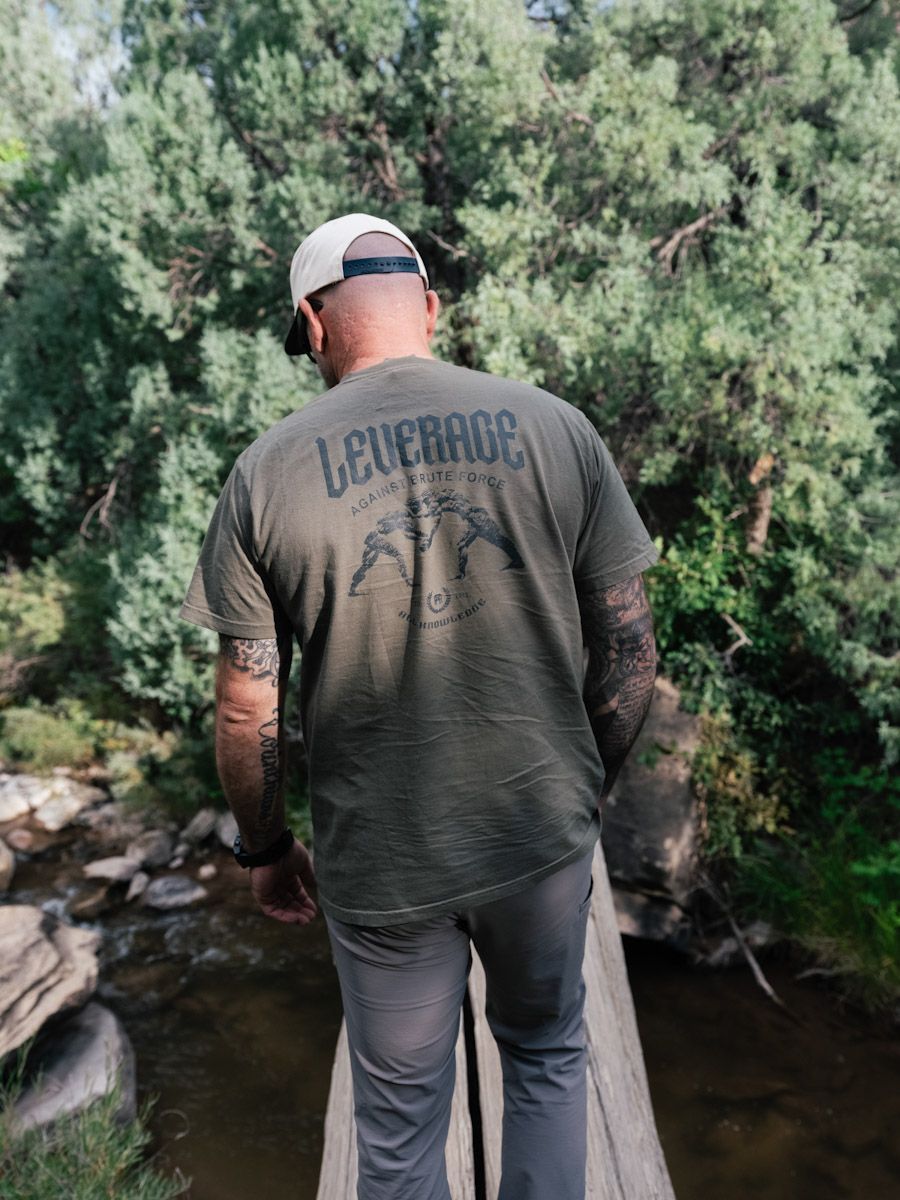 Man in green shirt, hat, and sunglasses, stands on a wooden bridge over a stream, surrounded by trees.