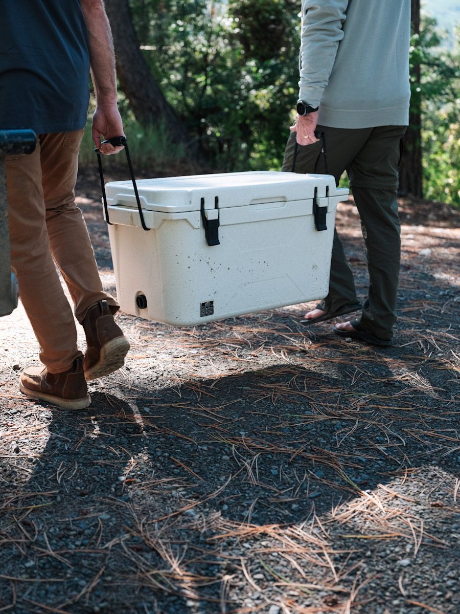 Two people pulling a large white cooler on a path in a forest.