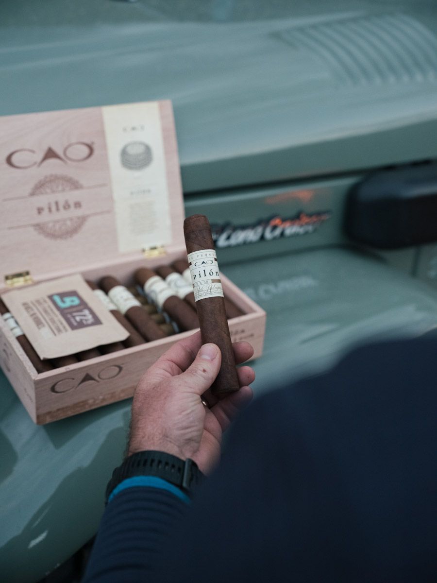 Man holding a dark cigar in front of a CAO box and a green Land Cruiser.