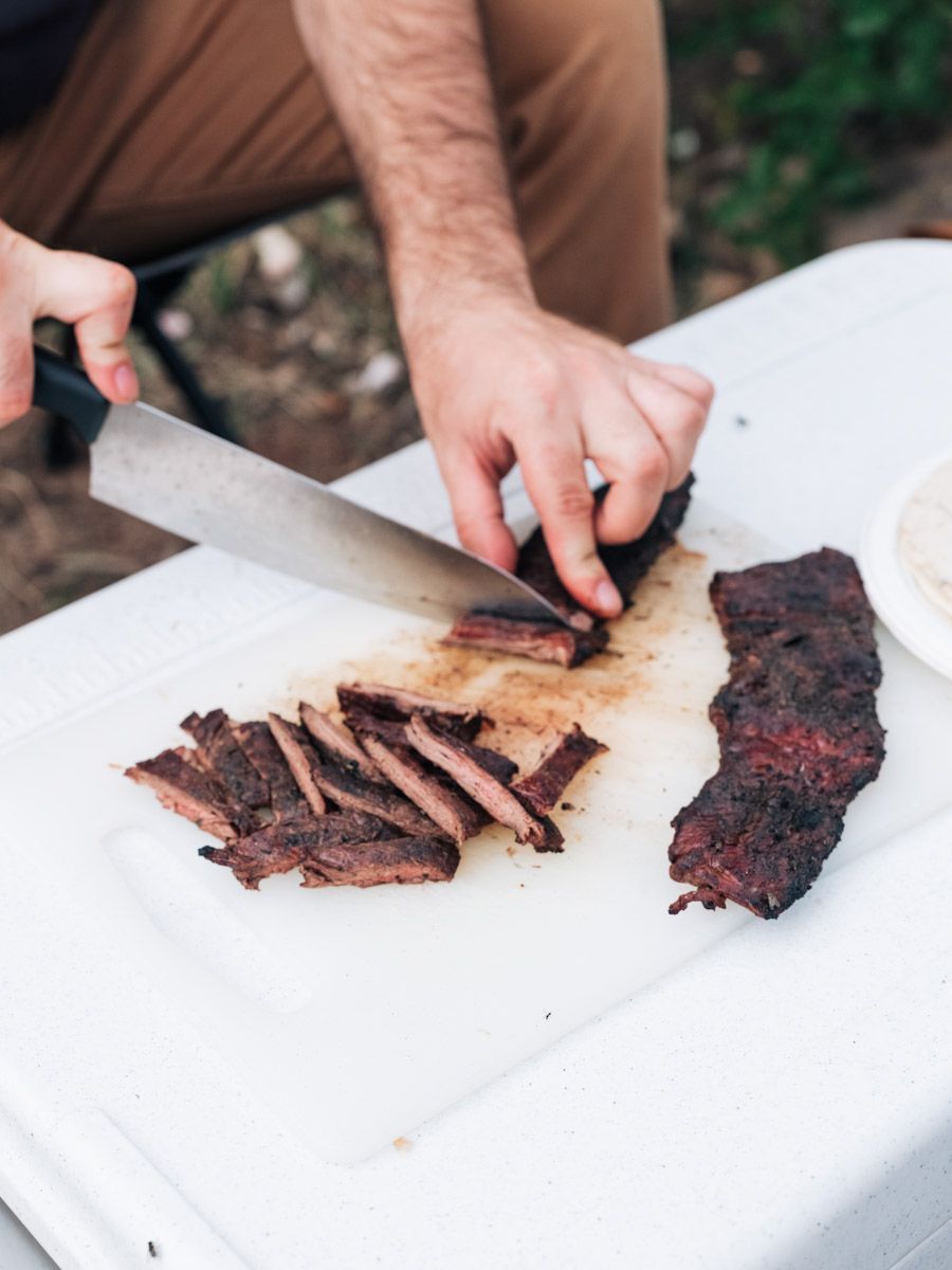 Person slicing cooked brisket on a white cutting board outdoors.