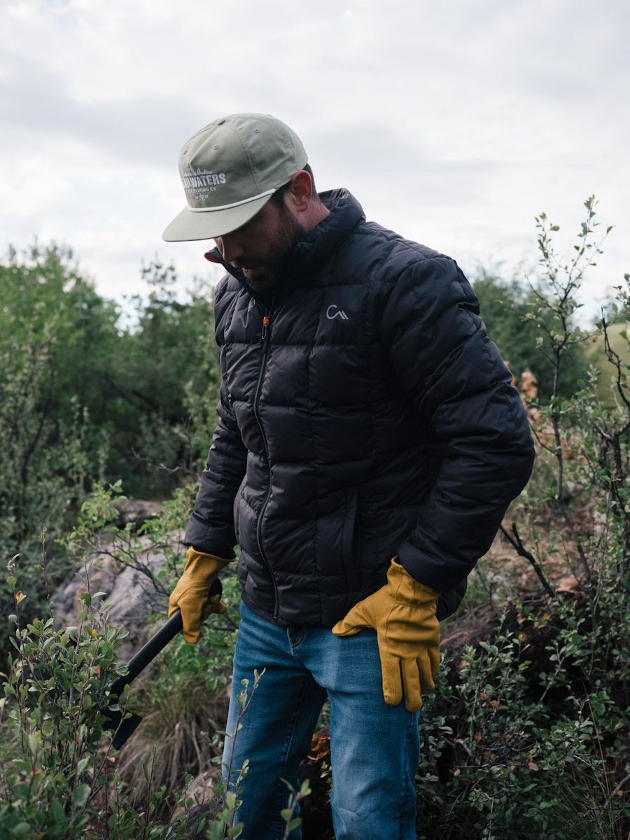 Man in black puffer jacket and gloves, working outdoors with a tool; wearing a cap, looking down.
