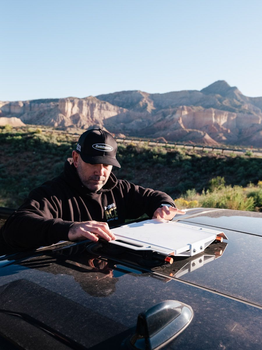 Man adjusting a white device on a car roof in front of a desert landscape.