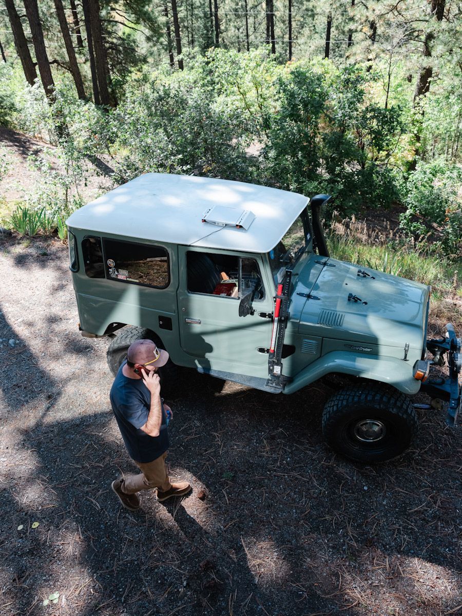 Man on phone stands near a green vintage 4x4. Forest background.