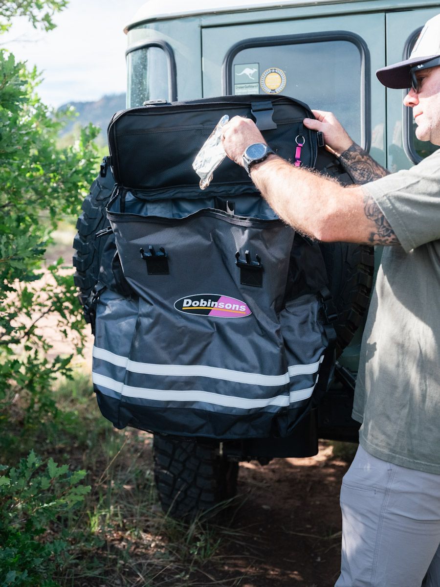 Man adjusts a black cargo bag mounted on the back of a green SUV with a spare tire.