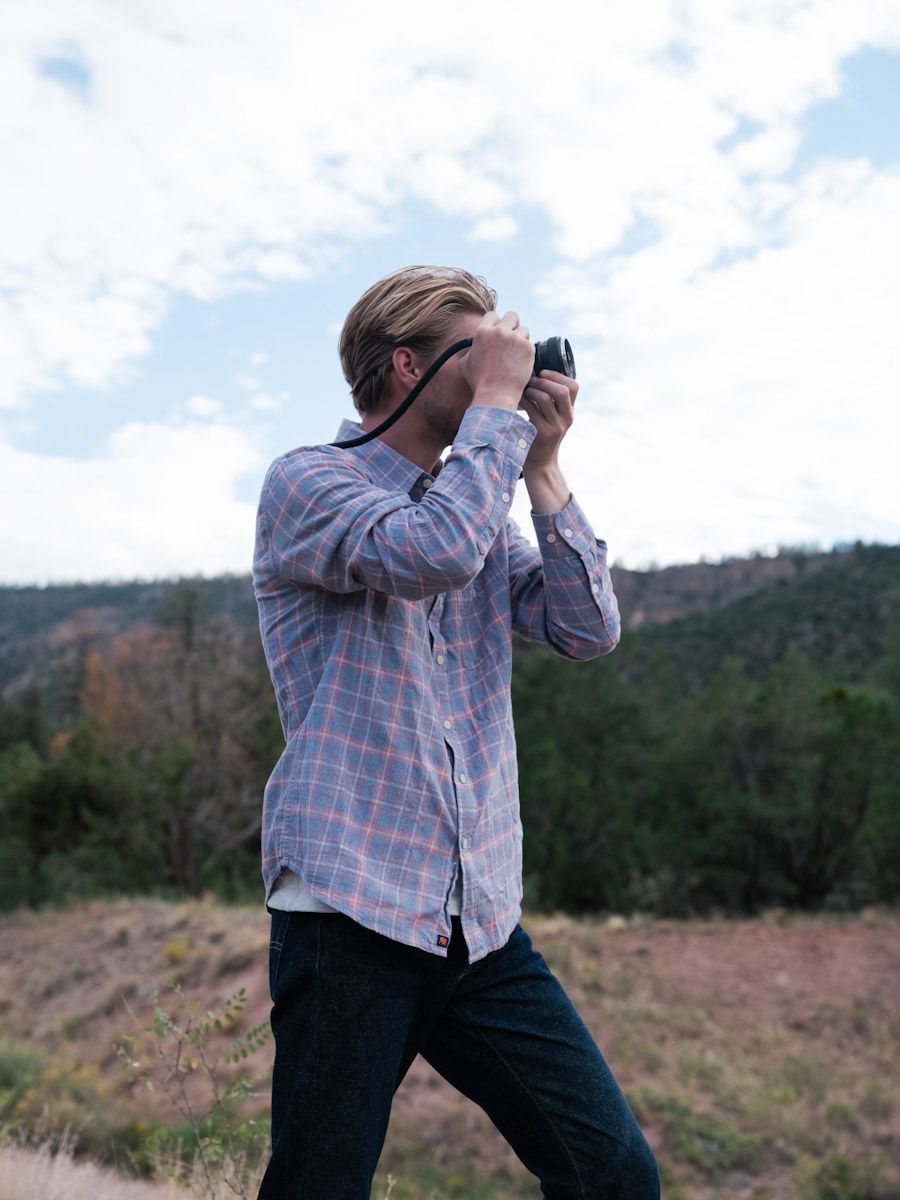 Blonde man in plaid shirt taking a photo with a camera, outdoors in a natural setting.