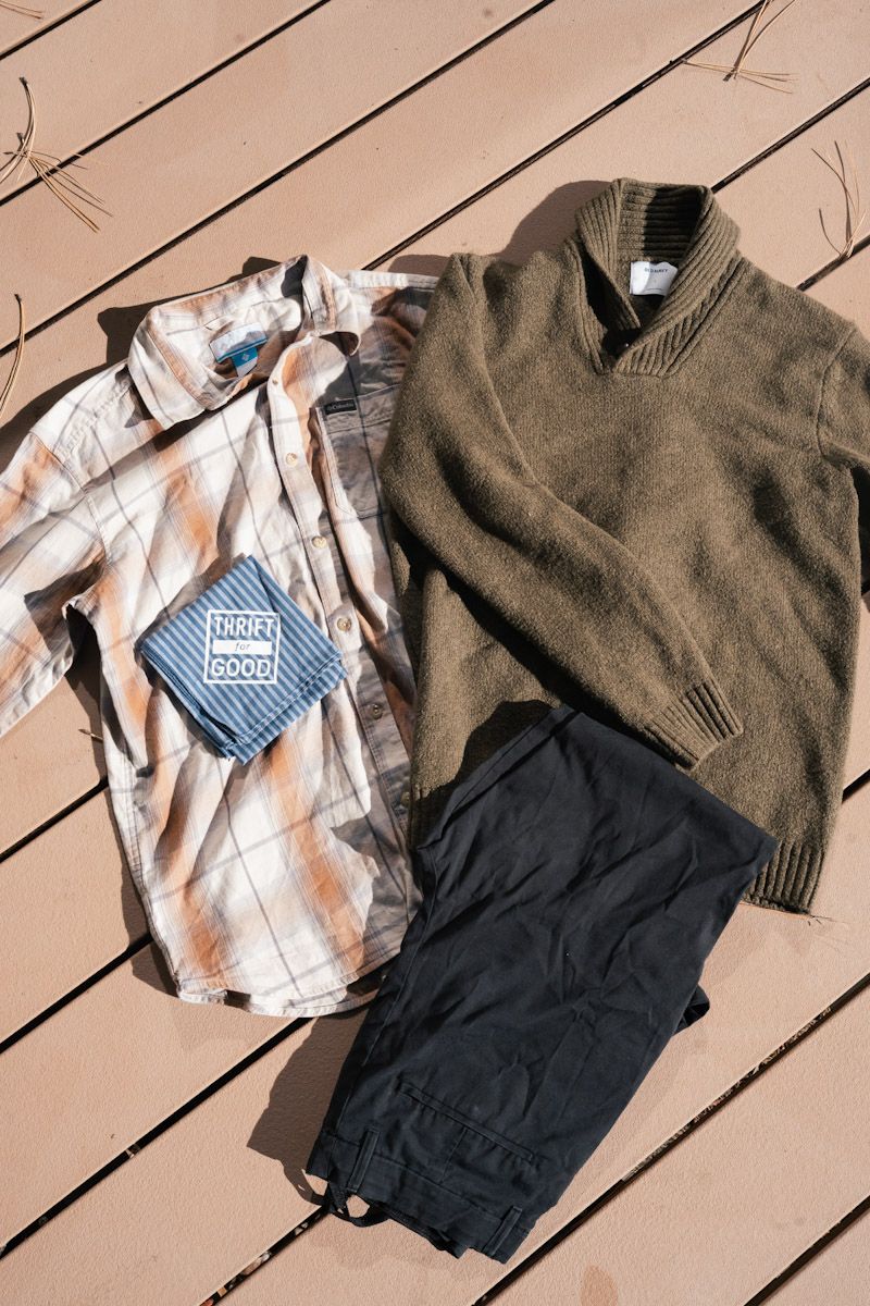 Men's clothing laid out on wooden planks: plaid shirt, green sweater, dark pants, and a blue handkerchief.