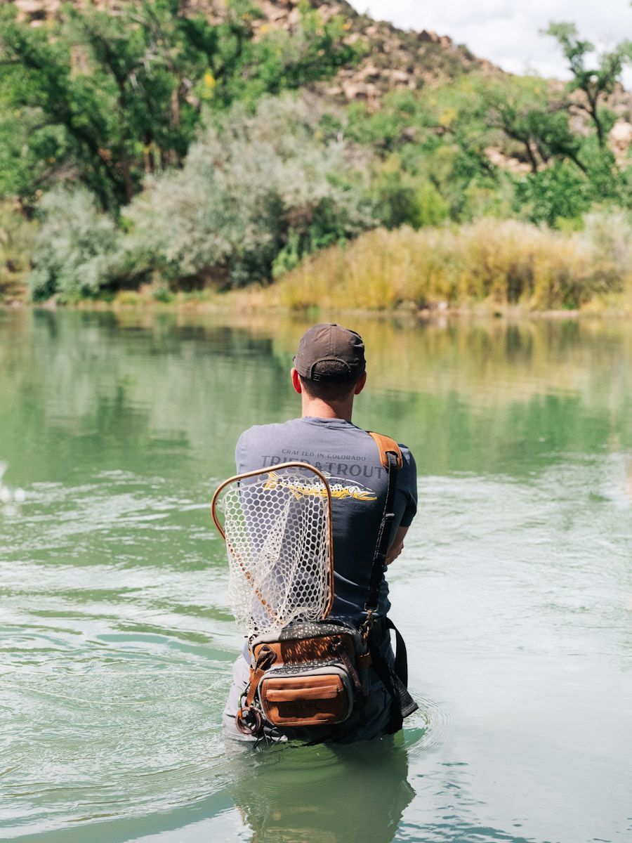 Person wading in river, wearing backpack with fishing net; green water and trees in background.