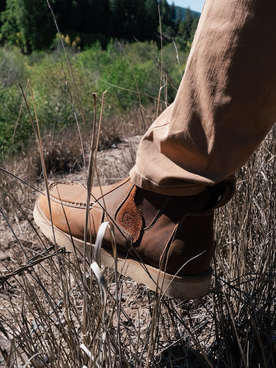 Brown boot stepping on dry grass in an outdoor setting. Tan pants visible.