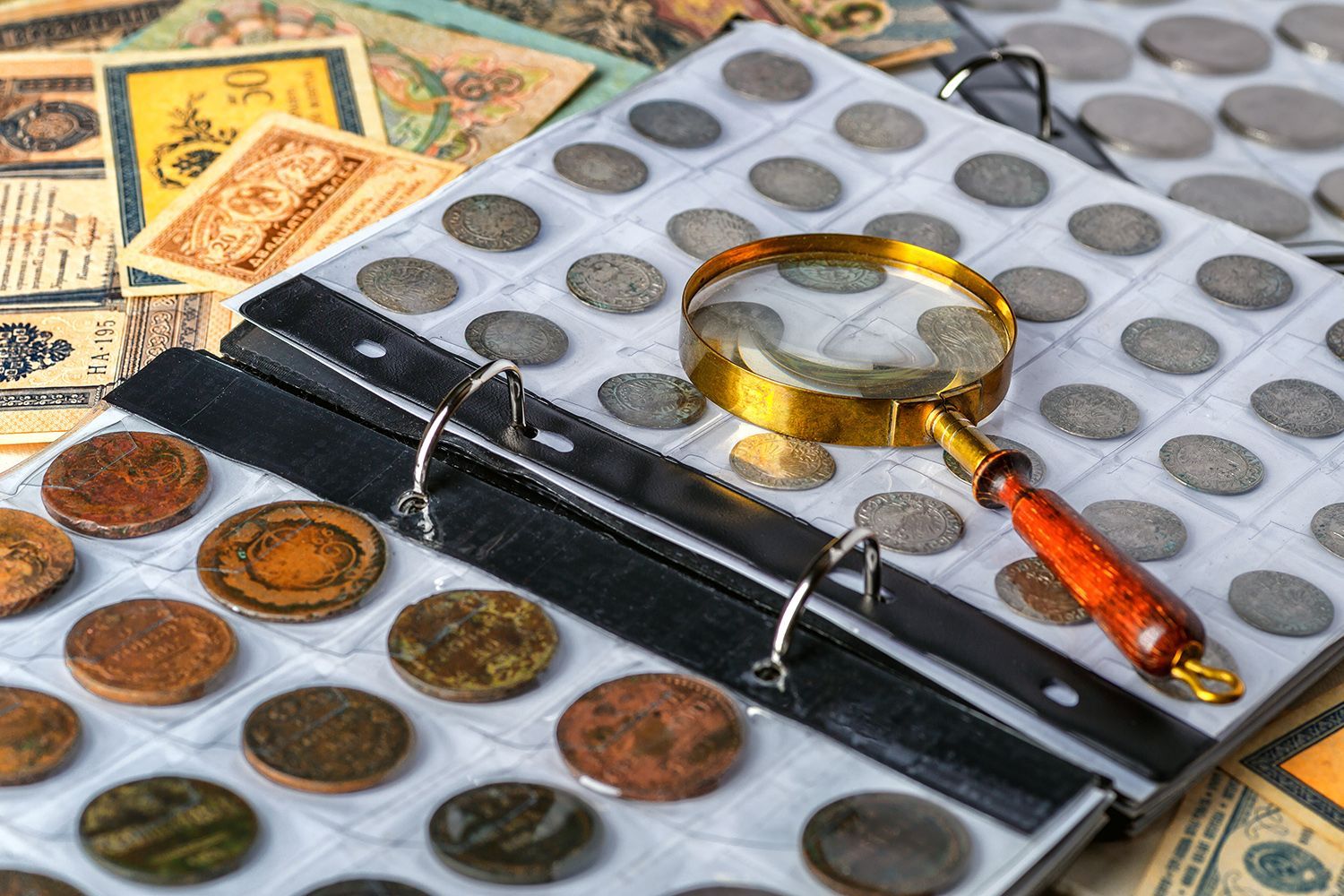Gold coin dealer examining rare coins album with magnifying glass and vintage currency notes.