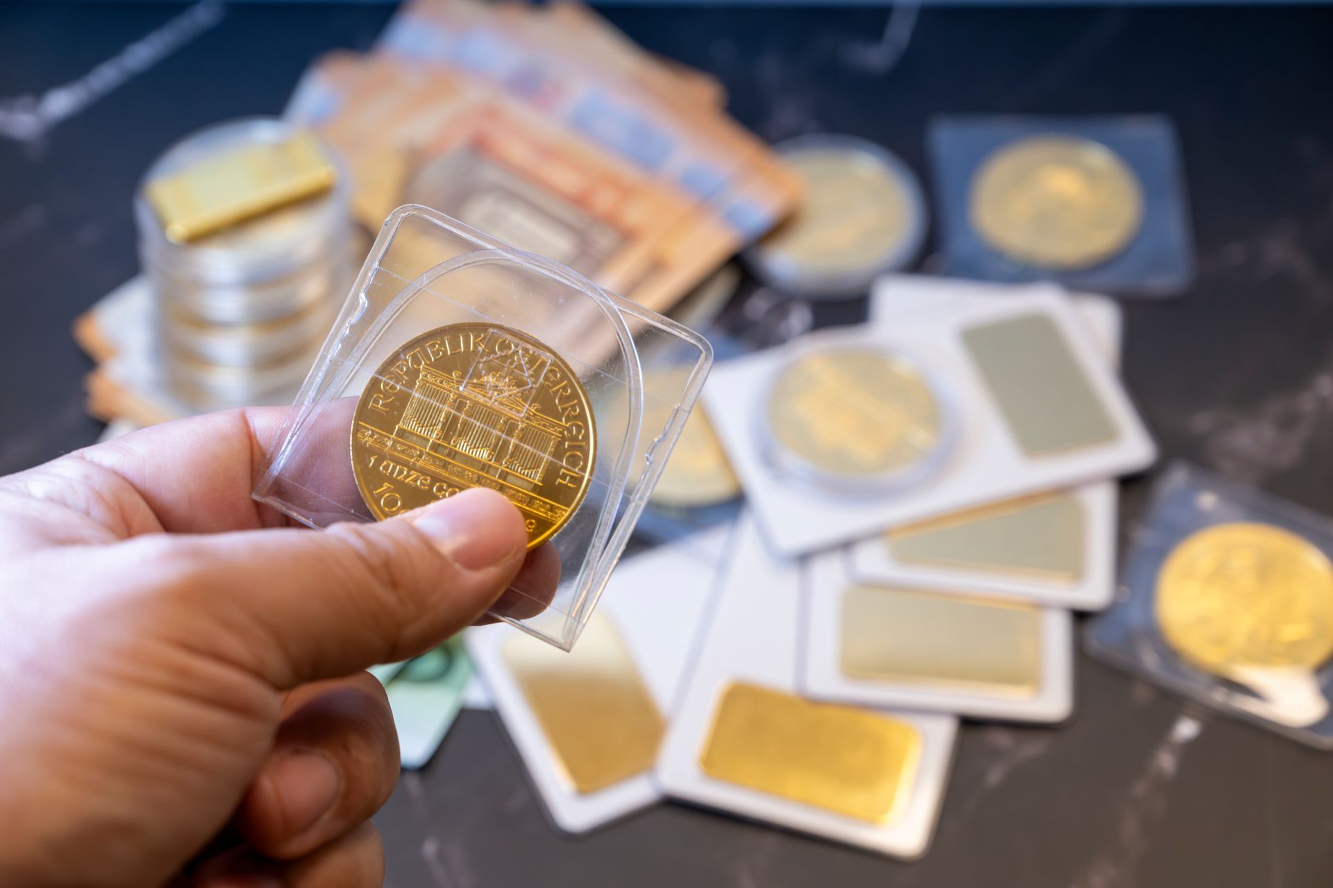 Hand holding a gold coin in a case with more coins and bars on a table.