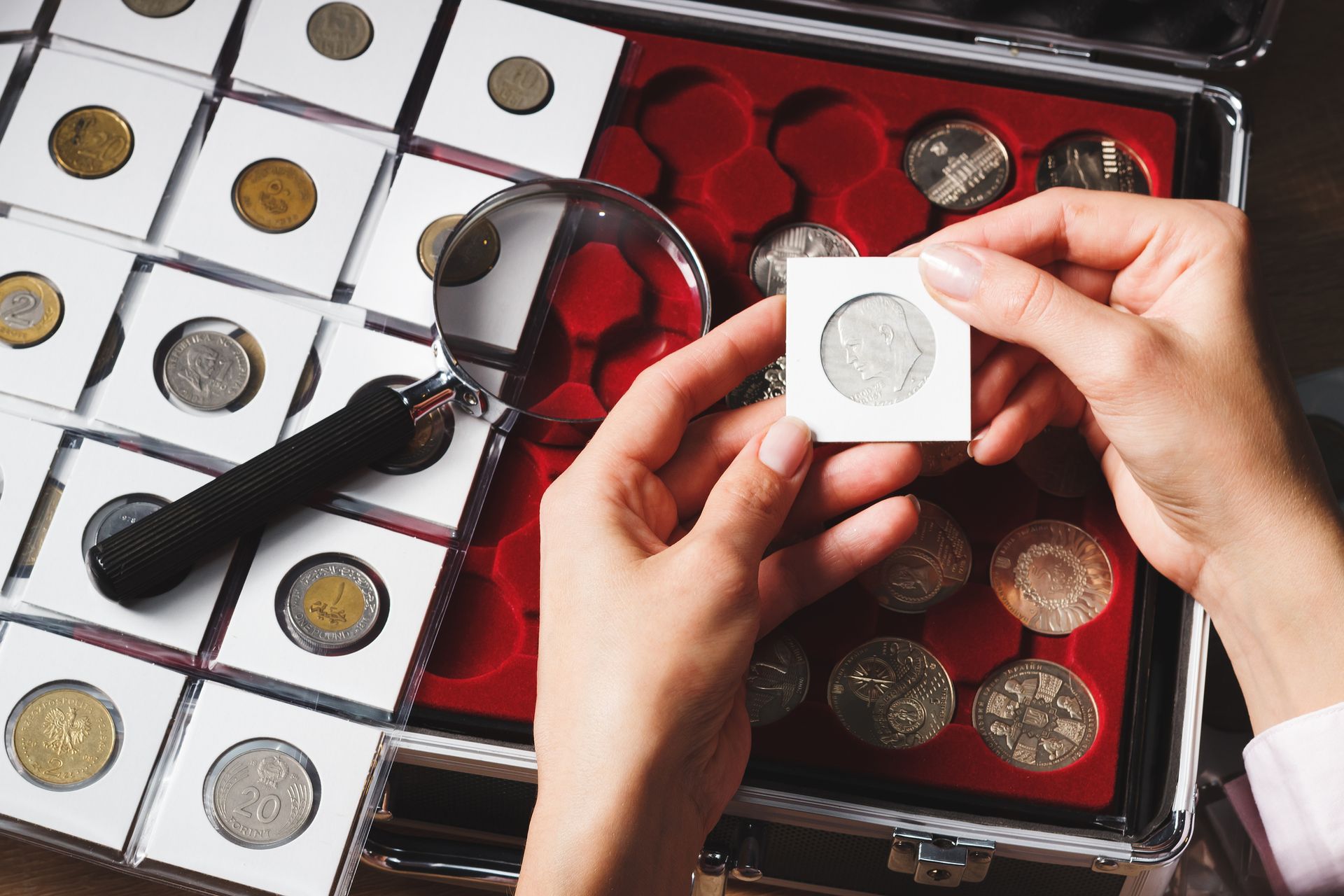 Hands inspect a coin in a protective case with a magnifier, surrounded by collectible coins.