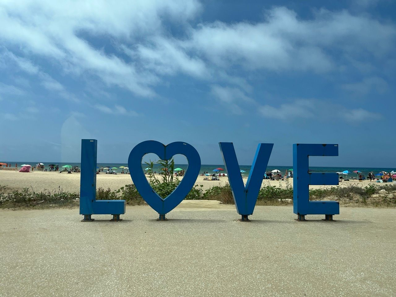 Blaues „LOVE“-Schild mit einem Herz in der Mitte, an einem Strand mit blauem Himmel und Wolken.