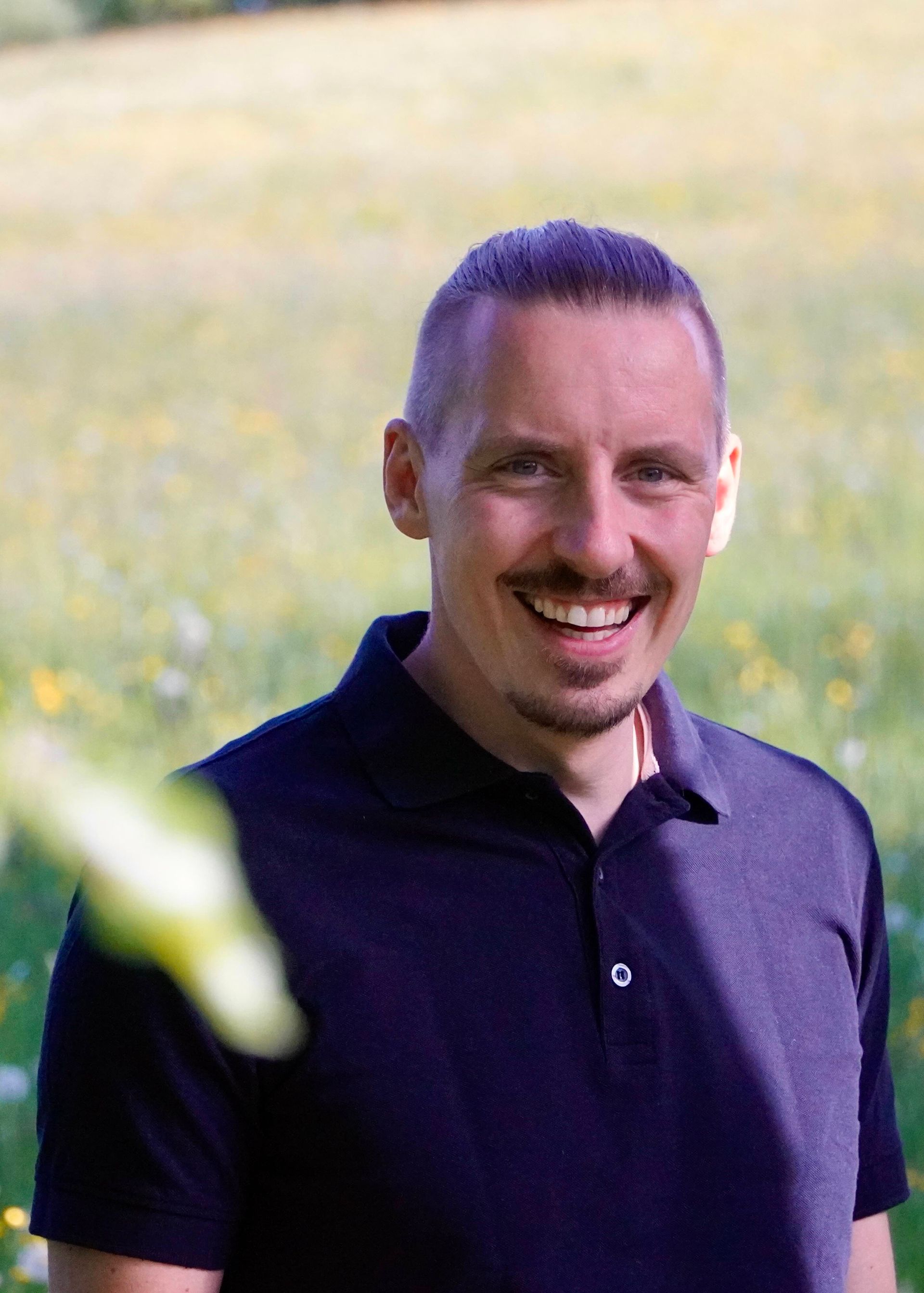 Mann mit kurzen Haaren, lächelnd auf einem Feld und schwarzem Poloshirt.