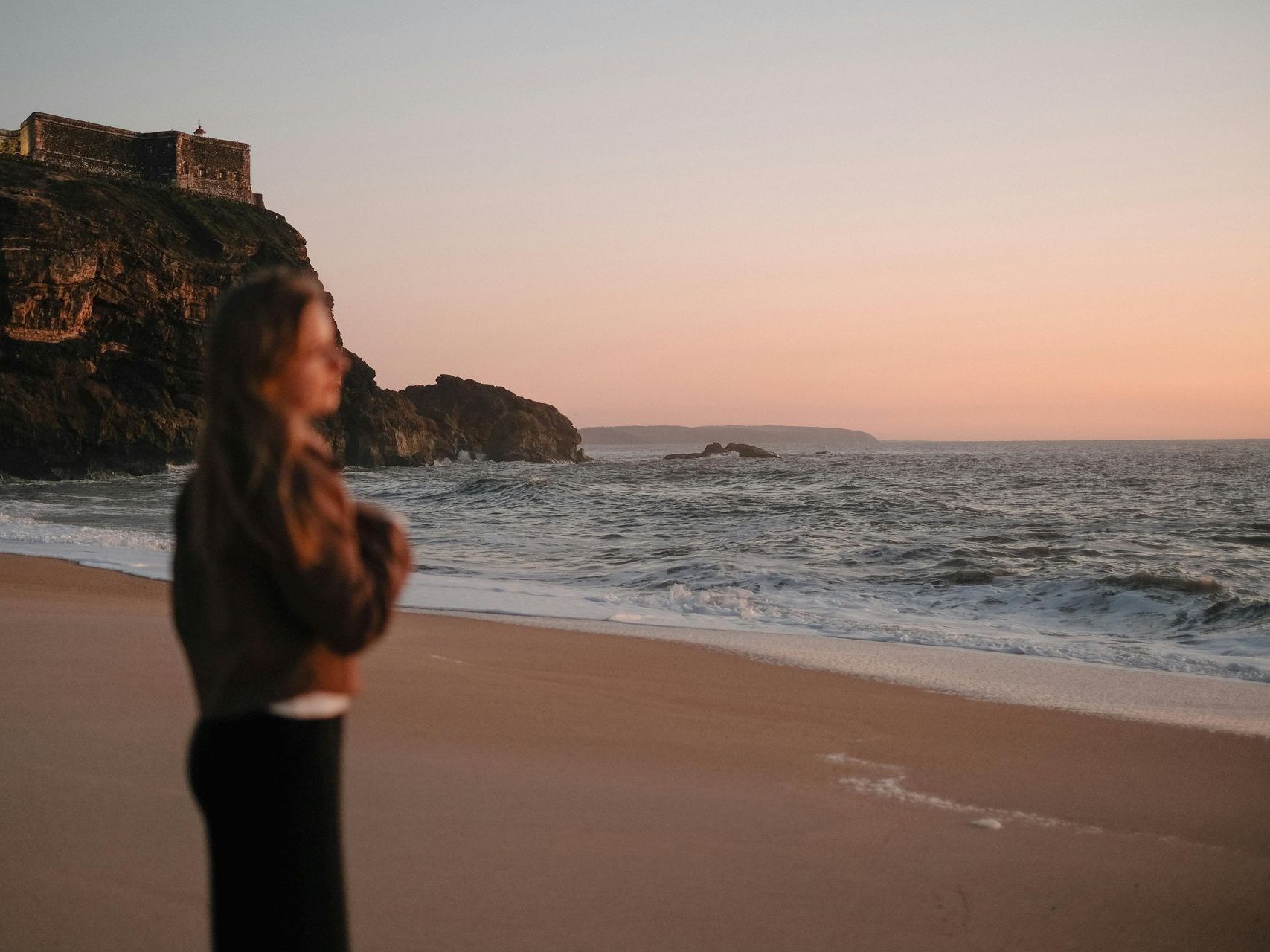 Frau am Strand blickt bei Sonnenuntergang auf das Meer. Im Hintergrund befindet sich eine Steinstruktur auf einer Klippe.