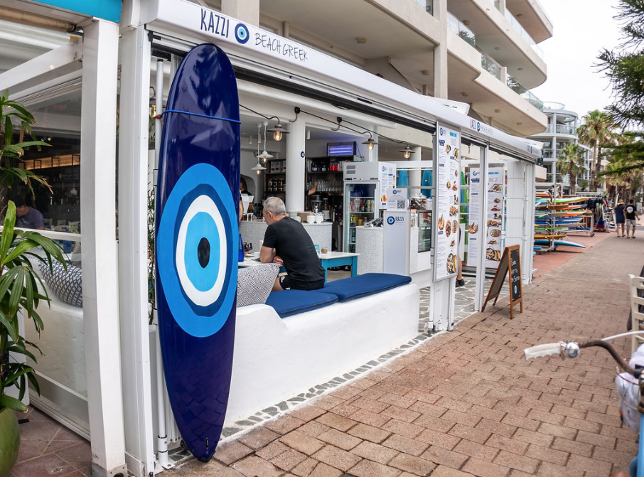 A man is sitting at a table in front of a restaurant with a blue evil eye on a surfboard.