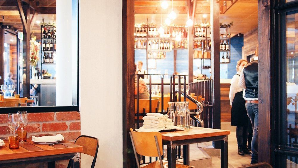 A restaurant with tables and chairs and people standing in the background.