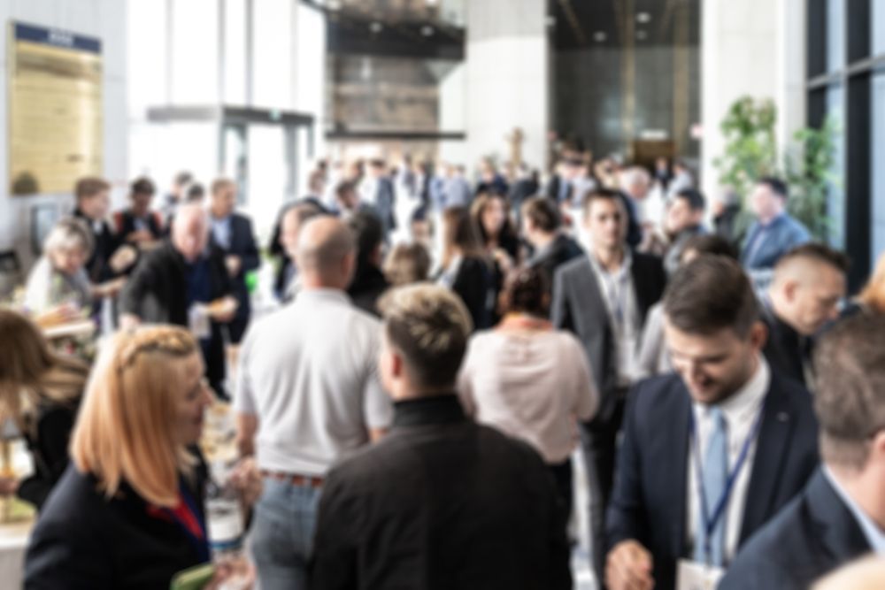 Crowded business networking event in a bright lobby, with people standing and talking in suits and name badges