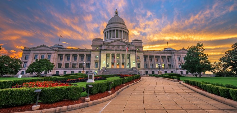 State capitol building at sunset with a domed center, landscaped gardens, and a curved walkway.