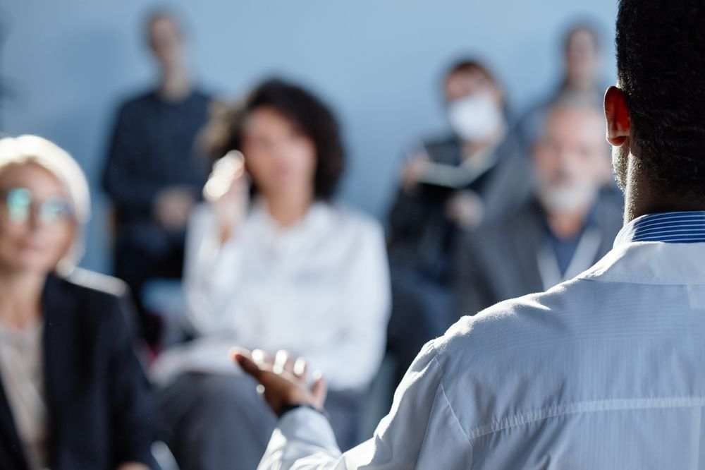 Speaker addressing an audience in a bright conference room, with attendees listening in the background