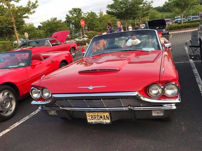 A bright red vintage Ford Thunderbird convertible parked in a lot during a sunny day car show.