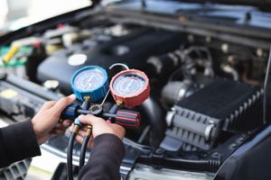 A technician holds a manifold gauge set while inspecting the air conditioning system of an open car engine bay.