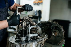 A mechanic in black gloves uses a wrench to work on the exposed metal gears of an automotive transmission.