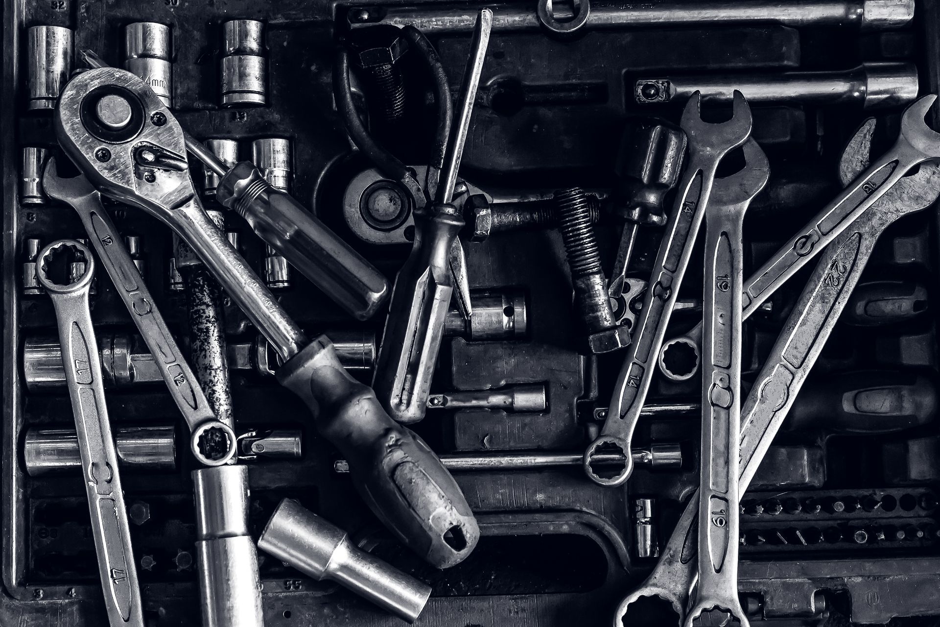 A monochromatic, top-down view of a cluttered tool box filled with various wrenches, a socket wrench, and assorted sockets.