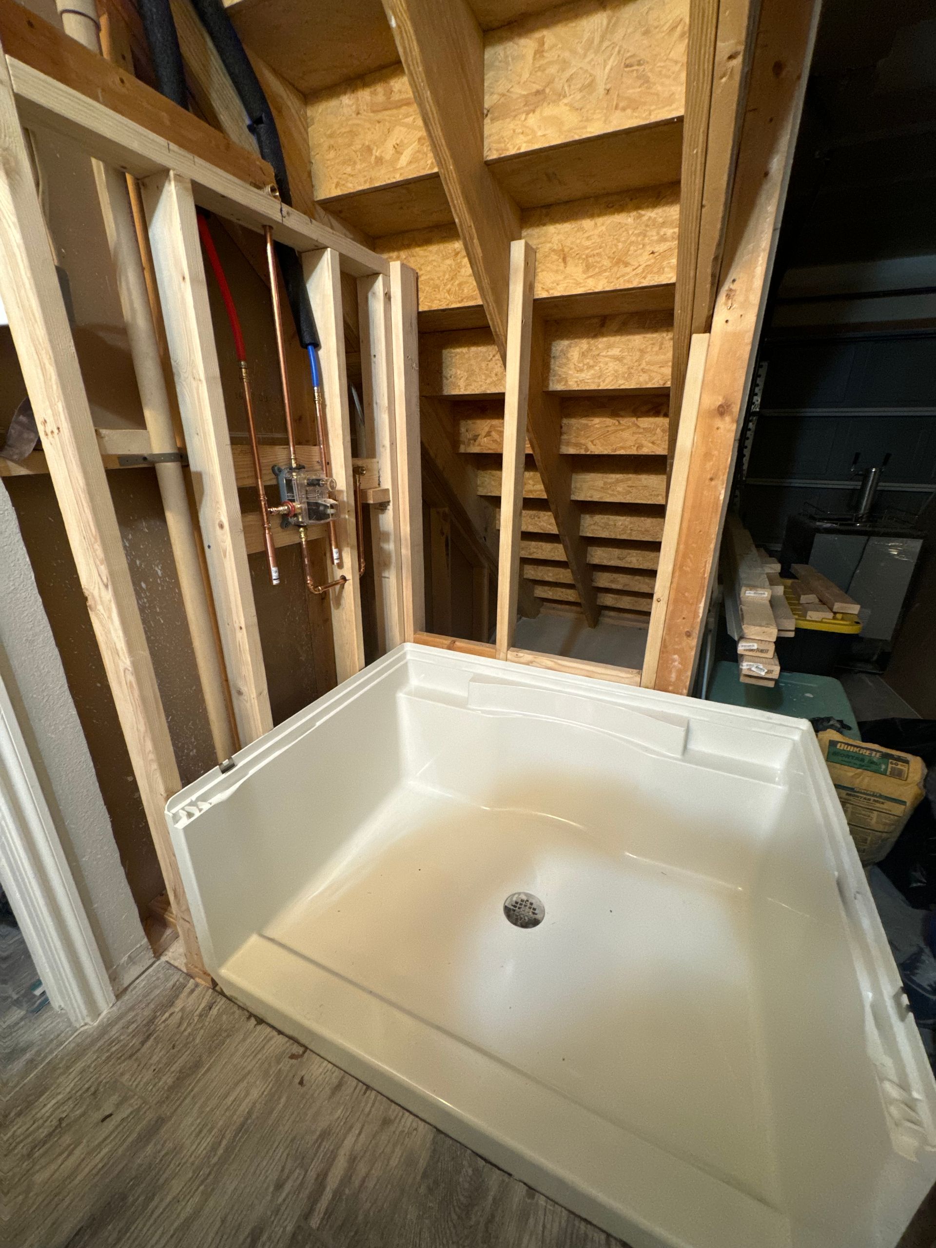 New shower base sits in a partially constructed bathroom; framing, plumbing visible. Staircase in background.