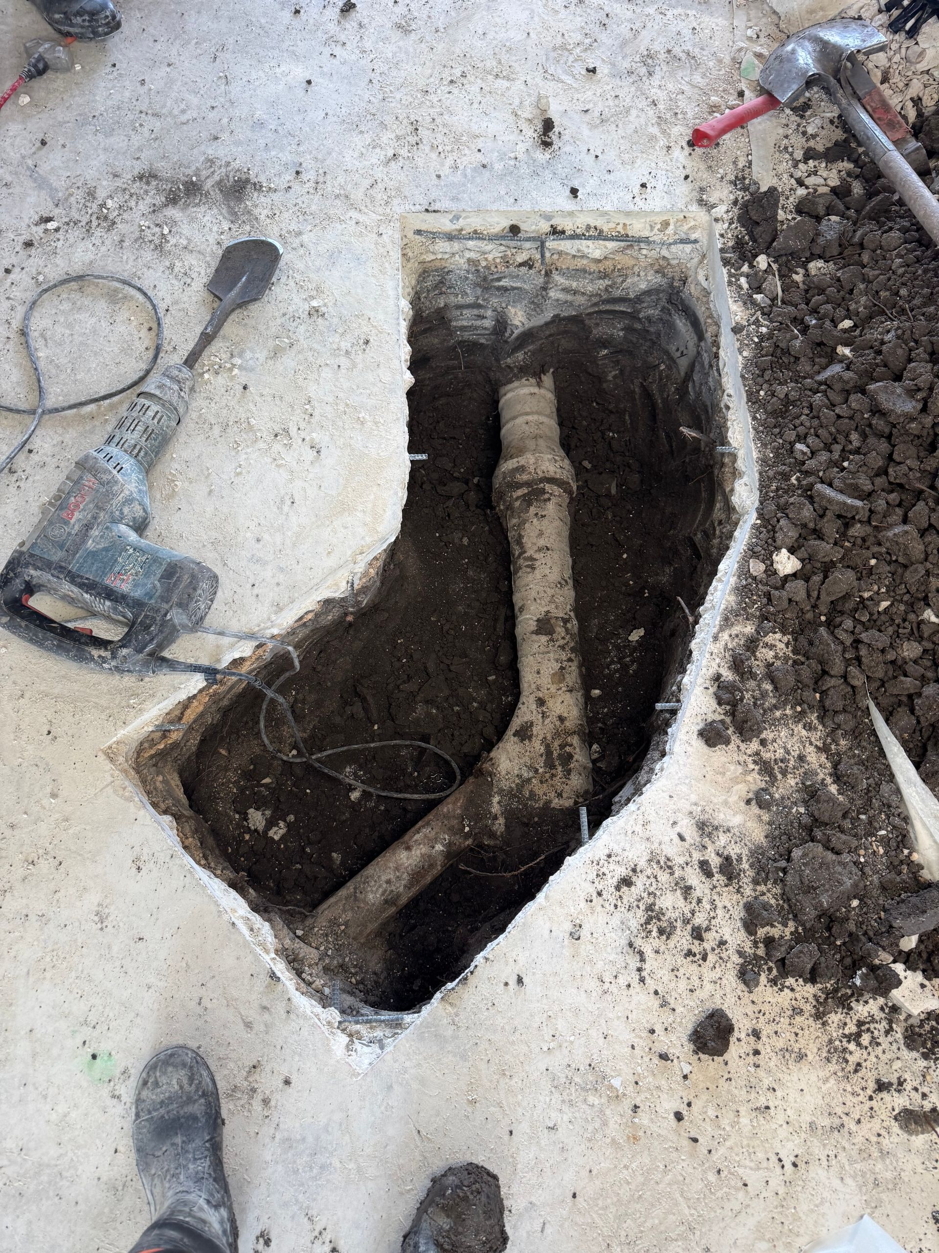 Hole dug in concrete floor exposing a curved pipe in dirt. Tools and boots surround the hole.