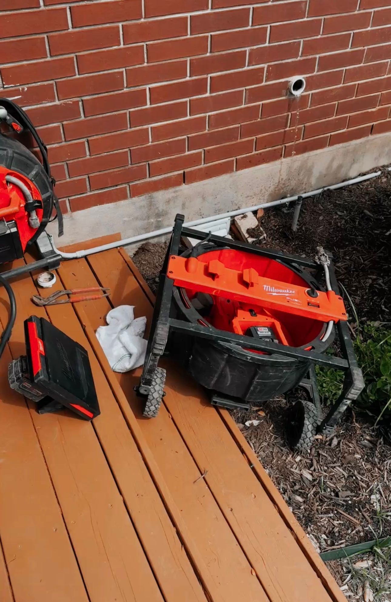 Red and black drain inspection equipment on a wooden deck next to a brick wall.