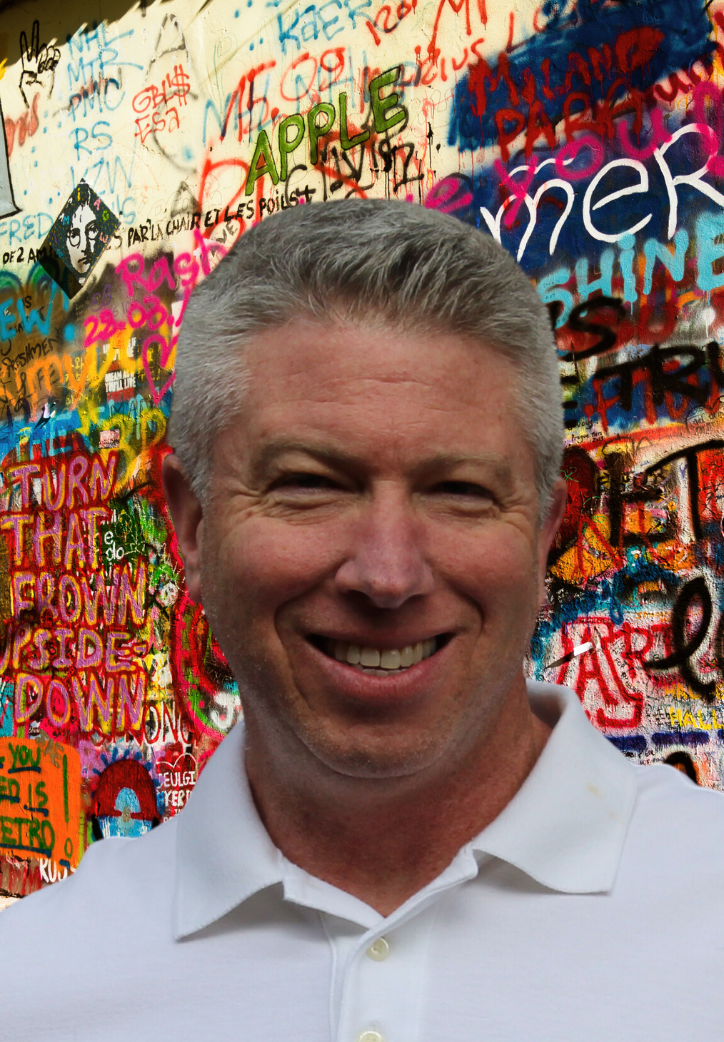 A man in a white shirt is smiling in front of a wall with graffiti on it