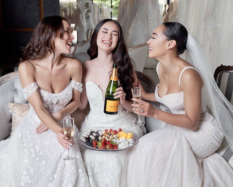 Three women in wedding dresses are sitting on a couch drinking champagne.